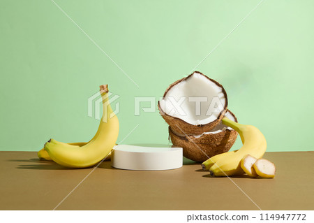 Advertising design template for products with fresh fruit ingredients such as bananas and coconuts against on green background with an empty showcase podium in the middle of brown table, frontal shot Advertising design template for products with fresh fruit ingredients such as bananas and coconuts against on green background with an empty showcase podium in the middle of brown table, frontal shot 114947772