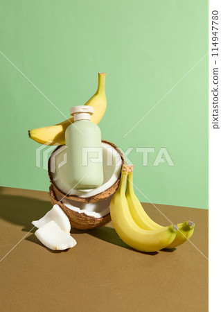 The green blank label bottle of cosmetic placed on a some coconuts piled on next to some fresh banana against light green background. Mock up for advertising with blank space for adding text 114947780