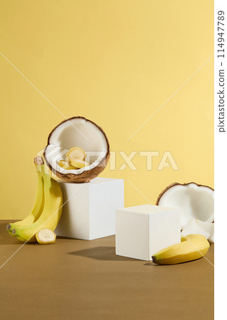 Creative vertical photo on yellow background, a rectangular empty platform has a half dried coconut above and a small bunch of bananas leaning on, next to an empty platform. Front view, copy space Creative vertical photo on yellow background, a rectangular empty platform has a half dried coconut above and a small bunch of bananas leaning on, next to an empty platform. Front view, copy space 114947789