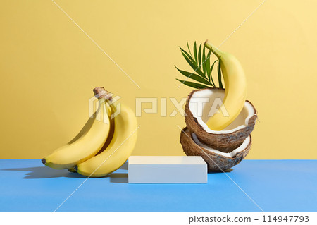 Tropical fruit concept photo with yellow background and blue countertop, an empty pedestal in rectangular shaped placed between a bunch of banana and a pile of dry coconut 114947793