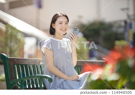 A young woman sitting on a bench in the city and looking at her smartphone 114948500