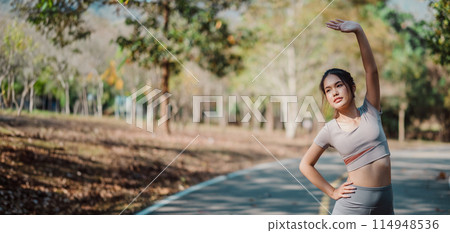 A woman is stretching on a road. She is wearing a grey shirt and black pants. The road is empty and there are no other people around 114948536