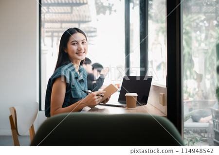 A woman is sitting at a table with a laptop and a notebook. She is smiling and she is enjoying her time. The scene suggests a relaxed and comfortable atmosphere. The woman might be working 114948542
