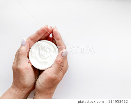 Hands middle age woman hold an open jar of white hand or body cream on white background with copy space. Flat lay composition. Skincare and beauty product concept. Top view. 114953412