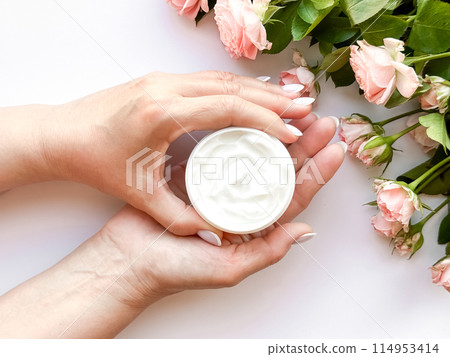 Hands middle age woman hold an open jar of white hand or body cream next to pink tea roses on white background, top view. Flat lay composition. Skincare and beauty product concept. Hands middle age woman hold an open jar of white hand or body cream next to pink tea roses on white background, top view. Flat lay composition. Skincare and beauty product concept. 114953414