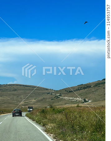 Winding road through mountain landscape, gentle distant mountains, sunny sky 114953757