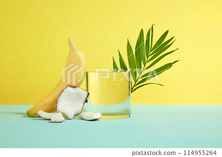 A empty glass podium placed in center with coconut pieces, banana and green coconut leaf decoration on blue table top over yellow background. Frontal shot photography with vacant space for design 114955264
