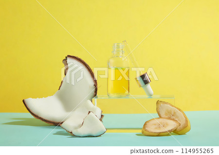 In the center of photo frame, a blank label serum bottle contains yellow essence displayed on a rectangular pedestal with some fresh banana and coconut decorated around against yellow background In the center of photo frame, a blank label serum bottle contains yellow essence displayed on a rectangular pedestal with some fresh banana and coconut decorated around against yellow background 114955265