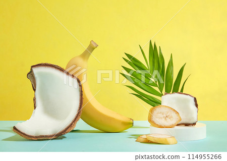 A yellow banana and a coconut piece leaning on each other, next to a round podium contains banana slices and coconut above, decorated with green coconut leaf behind. Copy space, front view 114955266