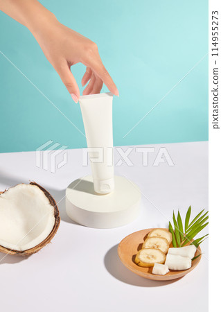 A beautiful female hand is lifting an unbranded cosmetic tube from round platform placed on white countertop, beside a half of dry coconut, a wooden tray of banana and coconut over blue background A beautiful female hand is lifting an unbranded cosmetic tube from round platform placed on white countertop, beside a half of dry coconut, a wooden tray of banana and coconut over blue background 114955273