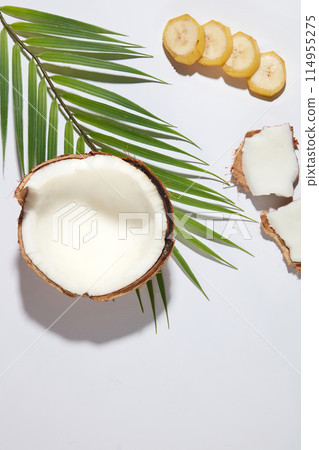 Top view photography at the white flat with banana and coconut flat lay above, banana slices arranged in a row next to some dried coconut pieces and a branch of coconut leaves decorated, blank space 114955275