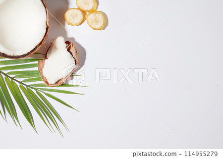 Half a coconut placed in the upper left corner of the photo with a white background, next to a few slices of banana and a branch of coconut leaves, remaining blank space to display organic products Half a coconut placed in the upper left corner of the photo with a white background, next to a few slices of banana and a branch of coconut leaves, remaining blank space to display organic products 114955279