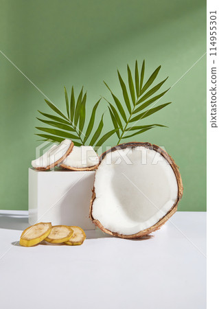 Photo was taken from front view on green background, a rectangular platform containing some pieces of coconut, next to a half of coconut and some banana slices, green coconut leaves decorated in back 114955301