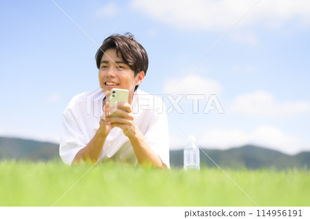 A man lying face down on the grass looking at his smartphone 114956191