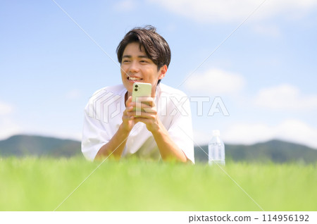 一名男子臉朝下躺在草坪上,看著他的智慧型手機 一名男子臉朝下躺在草坪上,看著他的智慧型手機 114956192