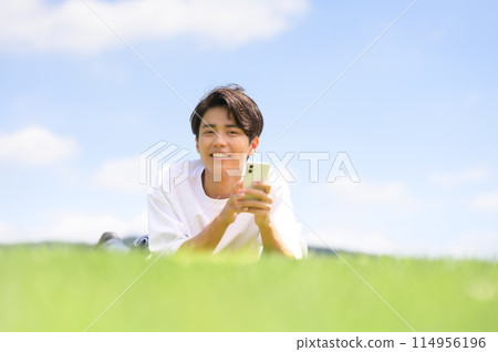 A man lying face down on the grass looking at his smartphone 114956196