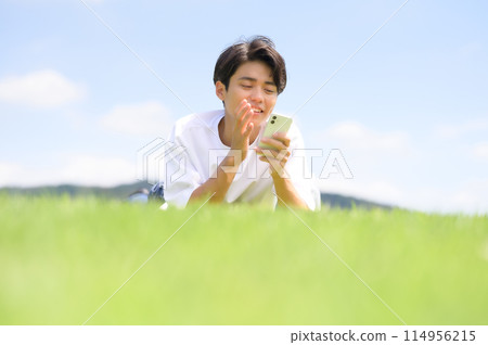 A man lying face down on the grass looking at his smartphone A man lying face down on the grass looking at his smartphone 114956215