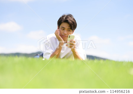 A man lying face down on the grass looking at his smartphone 114956216
