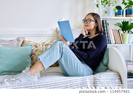 Young woman reading book, sitting on couch at home, engrossed in fiction 114957981