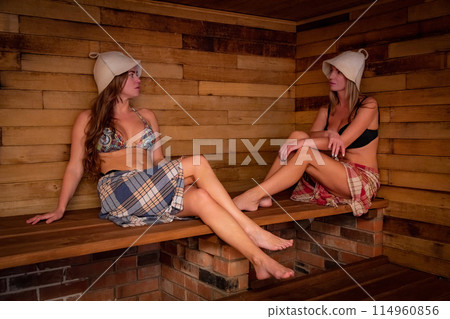 Two women in swimwear sitting on a wooden bench in a sauna Two women in swimwear sitting on a wooden bench in a sauna 114960856