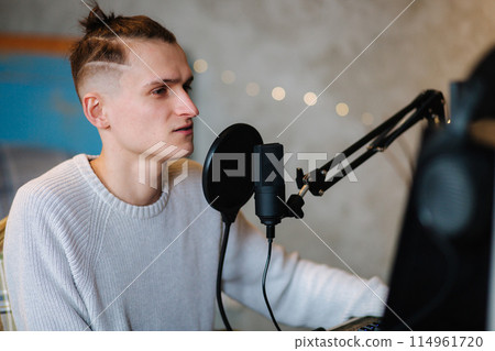 Young man makes a podcast audio recording at home. Man using pc and two professional microphones. Person recording a podcast at home using microphone and computers. Recording online podcast Young man makes a podcast audio recording at home. Man using pc and two professional microphones. Person recording a podcast at home using microphone and computers. Recording online podcast 114961720