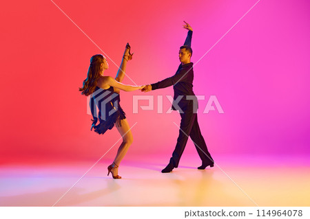 Professional ballroom dancers in black stages costumes performing in neon lighting against vibrant gradient background. 114964078