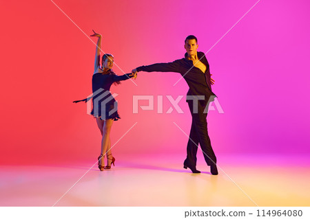 Talented ballroom dancers in black stages costumes dancing energetically in neon lighting against vibrant gradient background. 114964080