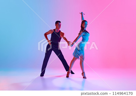 Ballroom dancers, man and woman in stage costumes gracefully performing in neon lighting against gradient background. Ballroom dancers, man and woman in stage costumes gracefully performing in neon lighting against gradient background. 114964170