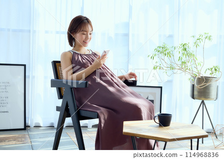 A woman sitting on a chair in a room and looking at her smartphone A woman sitting on a chair in a room and looking at her smartphone 114968836