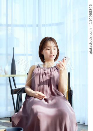 A woman sitting on a chair in a room and looking at her smartphone A woman sitting on a chair in a room and looking at her smartphone 114968846