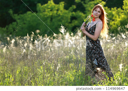 A girl with wild poppies enjoys the view from a hill near the river. Joy, happiness, serenity. Wild poppies by the river. 114969647