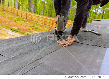 In roofer uses an air pneumatic nail gun to install new asphalt bitumen shingles In roofer uses an air pneumatic nail gun to install new asphalt bitumen shingles 114970159