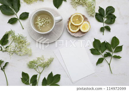 Cup of herbal elderflower drink, tea. Elderflowers, green leaves and fresh lemon fruit slices on old white wooden table. Blank greeting card, invitation mockup. Food floral pattern. Flat lay, top view 114972099