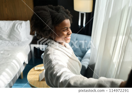 Side view portrait of happy young woman wearing bathrobe enjoying morning in hotel room and opening curtains 114972291