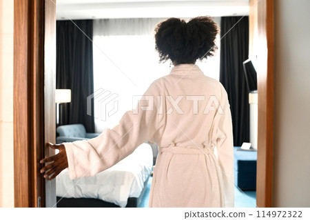 Back view of African American woman wearing bathrobe entering hotel room at resort, copy space 114972322