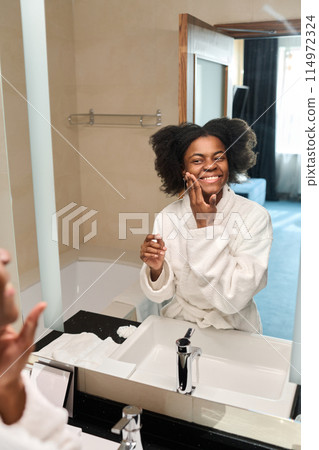 Vertical portrait of young African American woman looking in mirror enjoying skincare in morning, copy space 114972324