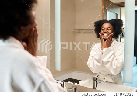 Portrait of smiling young Black woman looking in mirror enjoying skincare in morning, copy space 114972325