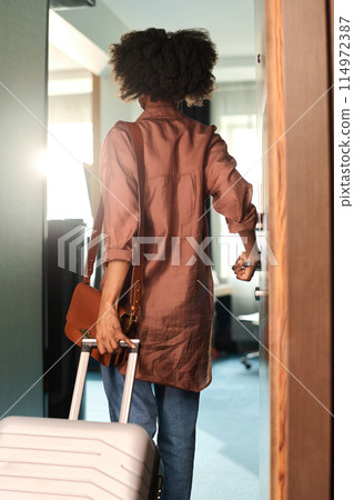 Vertical back view of young African American woman opening door to hotel room and holding suitcase 114972387