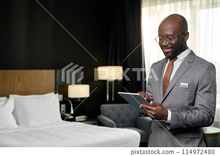 Waist up portrait of smiling African American man as hotel manager using digital tablet standing in hotel room copy space Waist up portrait of smiling African American man as hotel manager using digital tablet standing in hotel room copy space 114972480
