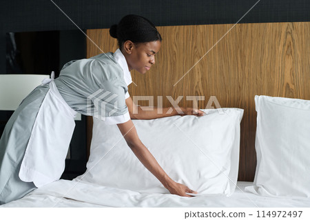 Side view portrait of young African American woman working as housekeeper in hotel and fluffing up fresh pillows copy space Side view portrait of young African American woman working as housekeeper in hotel and fluffing up fresh pillows copy space 114972497