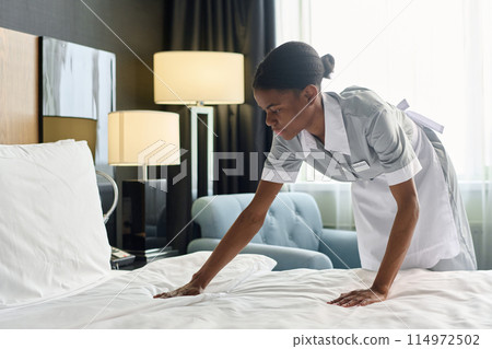 Side view portrait of young African American housekeeper working in hotel and making bed with fresh sheets copy space 114972502