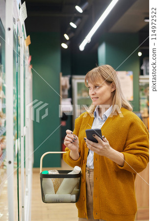 Vertical portrait of blonde adult woman grocery shopping in supermarket and using smartphone 114972522