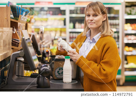 Waist up portrait of smiling blonde woman looking at camera in supermarket and using self checkout machine copy space Waist up portrait of smiling blonde woman looking at camera in supermarket and using self checkout machine copy space 114972791