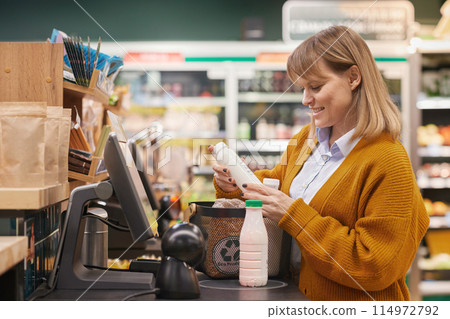 Side view portrait of smiling Caucasian woman using self checkout machine buying milk and groceries in supermarket Side view portrait of smiling Caucasian woman using self checkout machine buying milk and groceries in supermarket 114972792