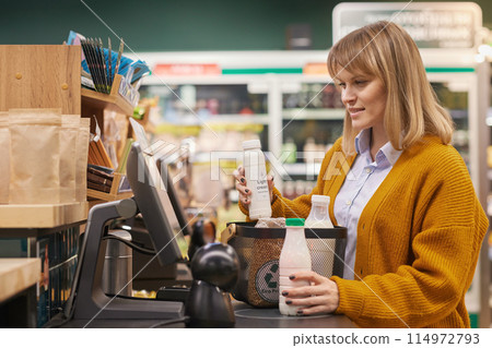 Side view portrait of blonde adult woman using self checkout machine while buying dairy products in supermarket and smiling Side view portrait of blonde adult woman using self checkout machine while buying dairy products in supermarket and smiling 114972793