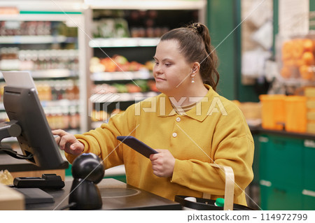 Side view portrait of smiling woman with Down syndrome using self checkout machine in supermarket and holding smartphone 114972799