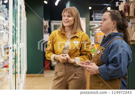 Portrait of smiling manager training young woman with Down syndrome working in supermarket and looking at freezer display Portrait of smiling manager training young woman with Down syndrome working in supermarket and looking at freezer display 114972843