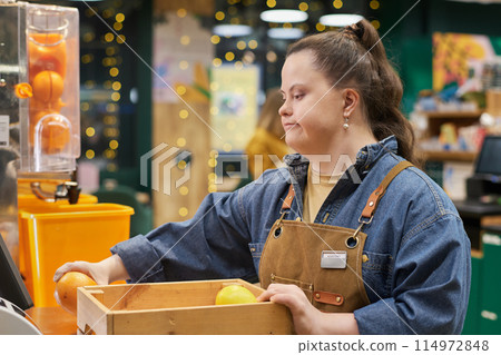 Side view portrait of smiling young woman with disability working at fresh juice stand in supermarket and sorting oranges Side view portrait of smiling young woman with disability working at fresh juice stand in supermarket and sorting oranges 114972848