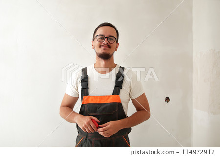 Construction worker in overalls indoor. Smiling young man on white wall background, holding tape measure. cheerful builder with tape measure 114972912