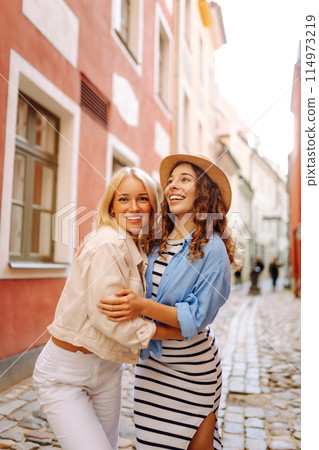 Two women are walking down a street, one wearing a white jacket and the other wearing a blue shirt and white skirt. They are smiling and holding hands, giving off a happy and friendly vibe 114973219